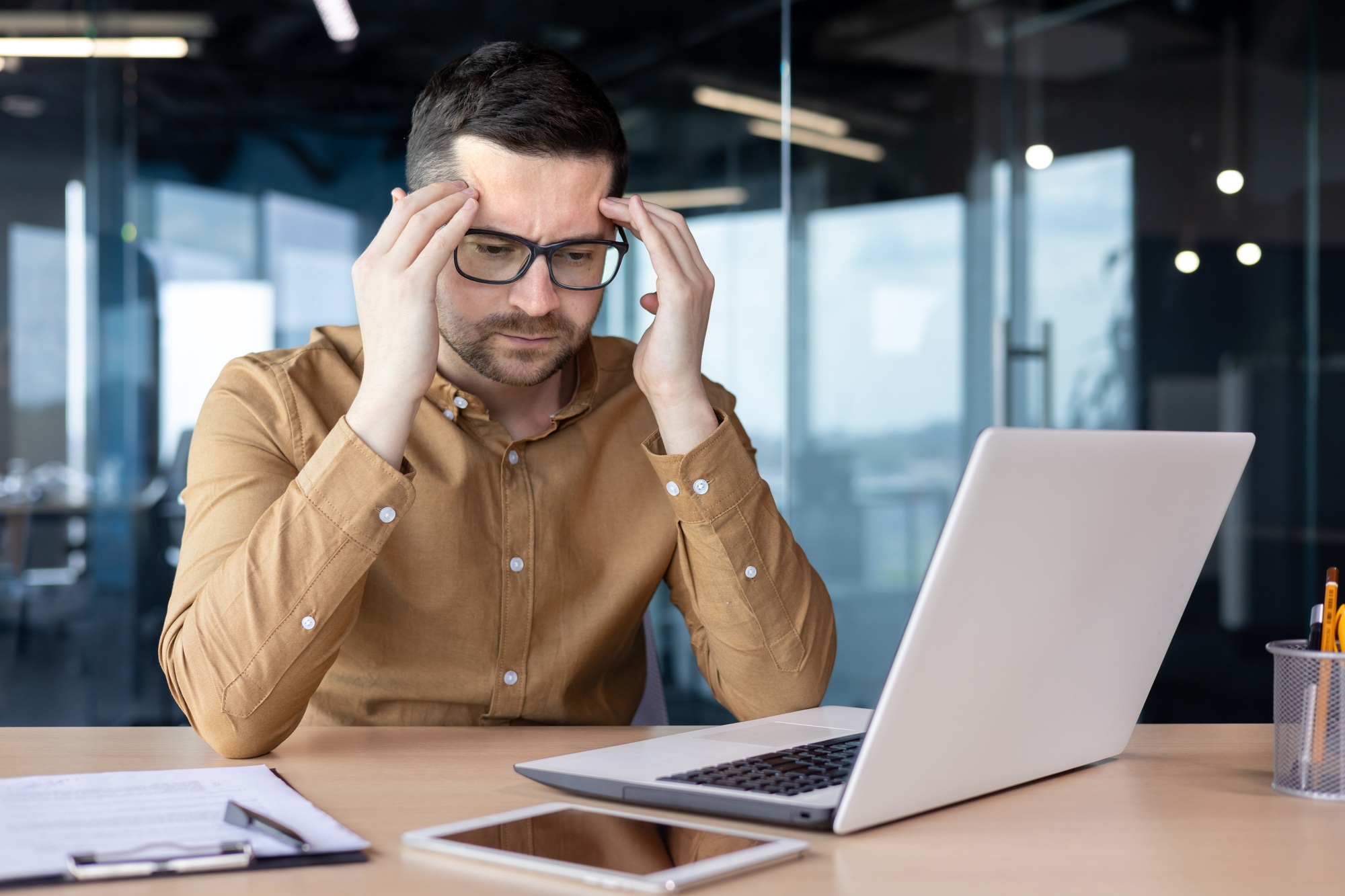 A worried young male businessman sits at a desk in the office in front of a laptop and holds his